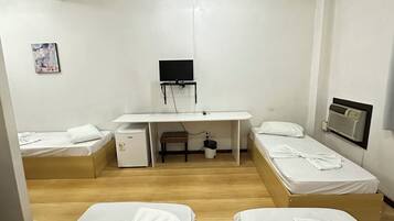 A hotel room with two single beds, a desk with a flat-screen TV, a small table, and a wall-mounted air conditioner.