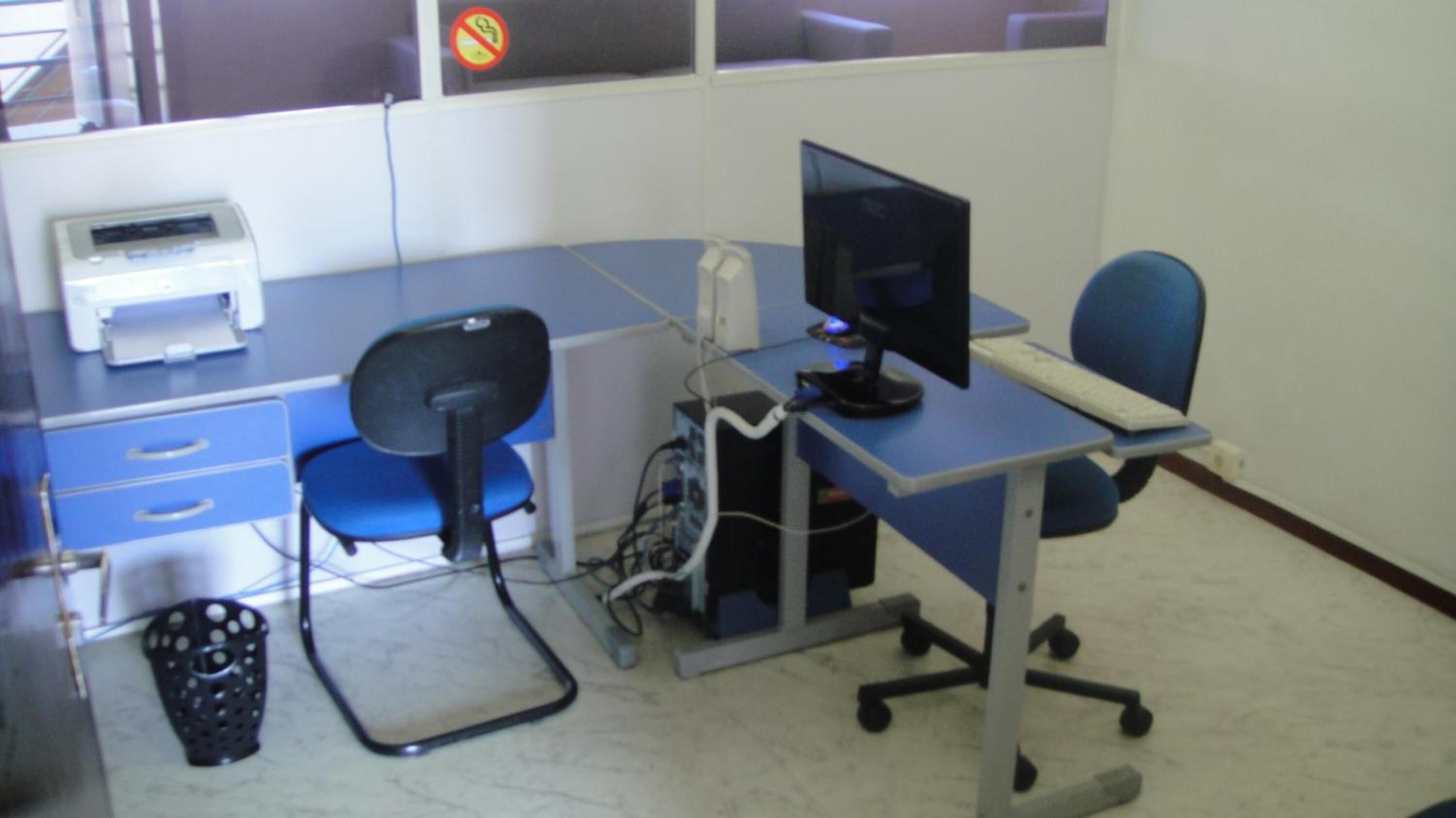 Uma escrivaninha com monitor, teclado e impressora. Há uma cadeira de escritório azul e uma lixeira.