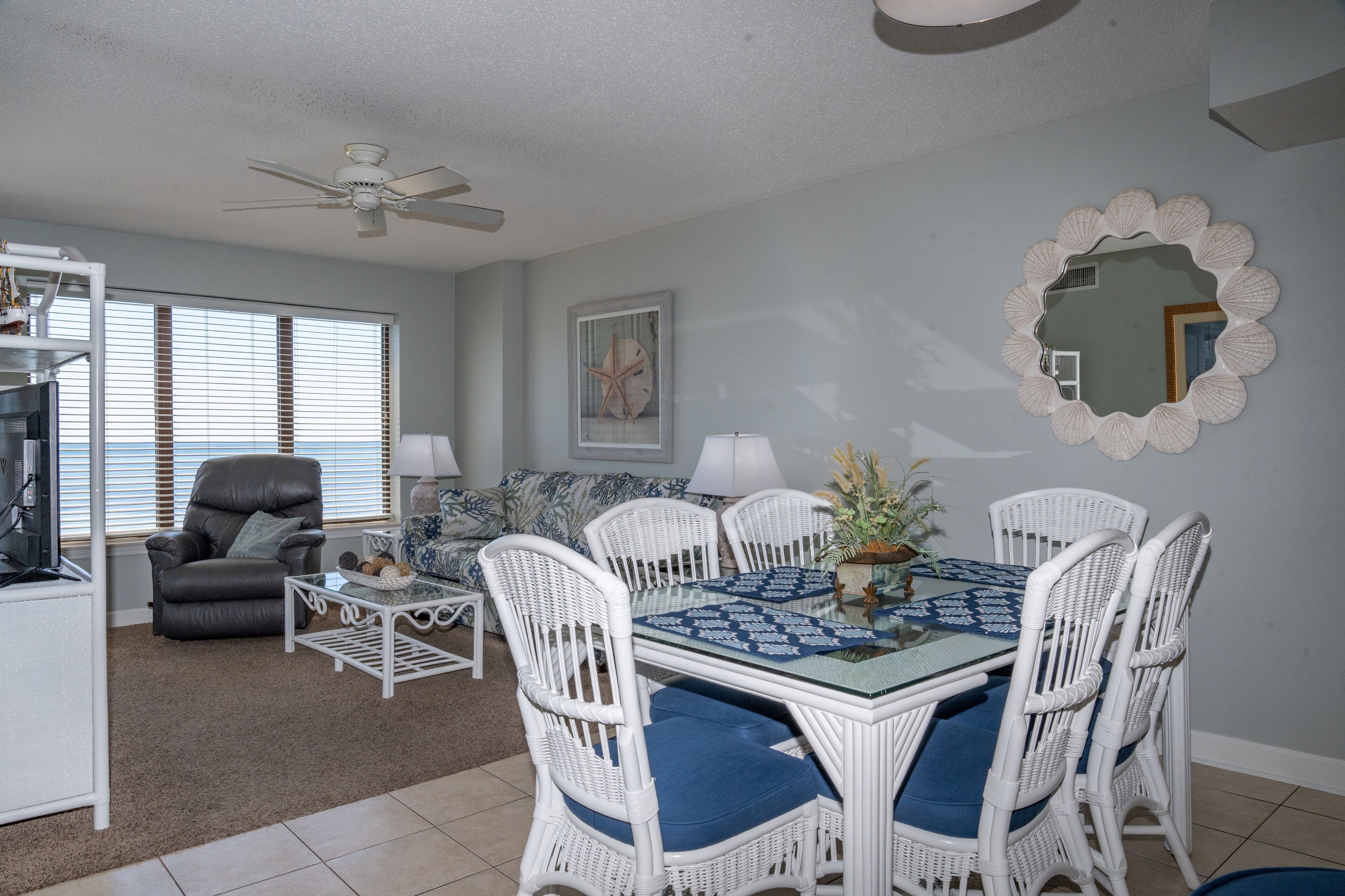 A dining area with a glass-top table, white wicker chairs with blue cushions, and a decorative mirror on the wall.