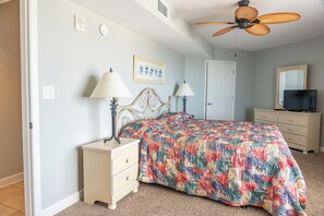 A bedroom with a bed, two bedside tables with lamps, a ceiling fan, a door, a dresser, and a television.