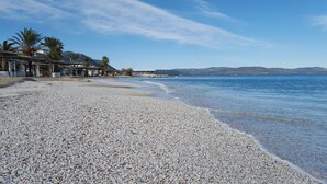A pebbled beach with a clear blue sea, palm trees, and a distant mountain range.