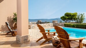 A poolside area with wooden chairs and a table, a stone wall, and a view of the sea.