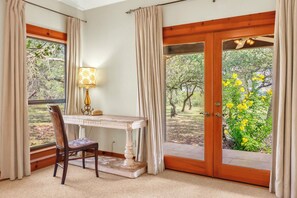 A room with a desk, chair, and a view of greenery through a door.