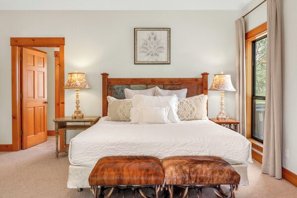 A bedroom with a large bed, two bedside tables, a wooden headboard, a framed picture on the wall, and a window with curtains.