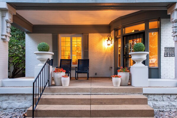 A building entrance with a set of steps leading up to a door, flanked by two large planters with flowers, a bench, and a wall-mounted light fixture.