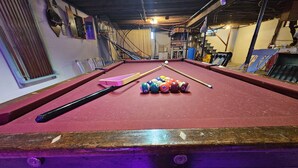 Une table de billard avec des billes et des queues, entourée d’étagères et d’un escalier.