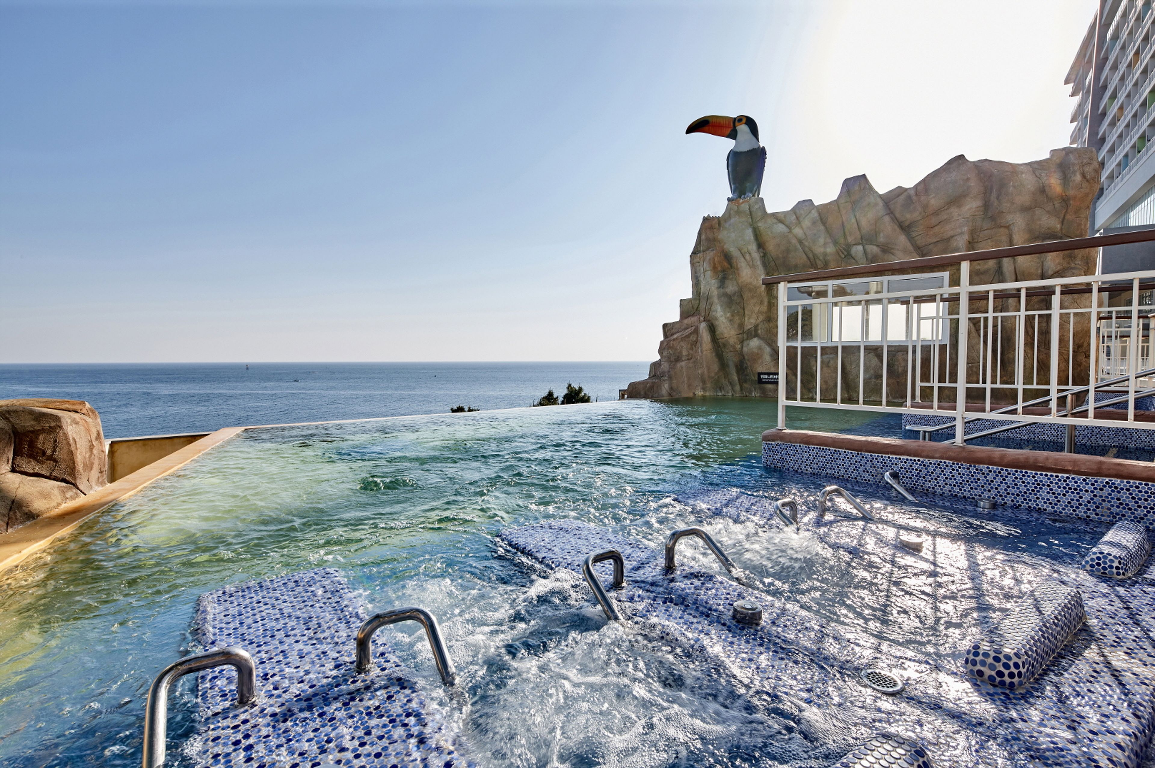 A pool area with a view of the ocean, a rocky outcrop, and a building with a balcony.