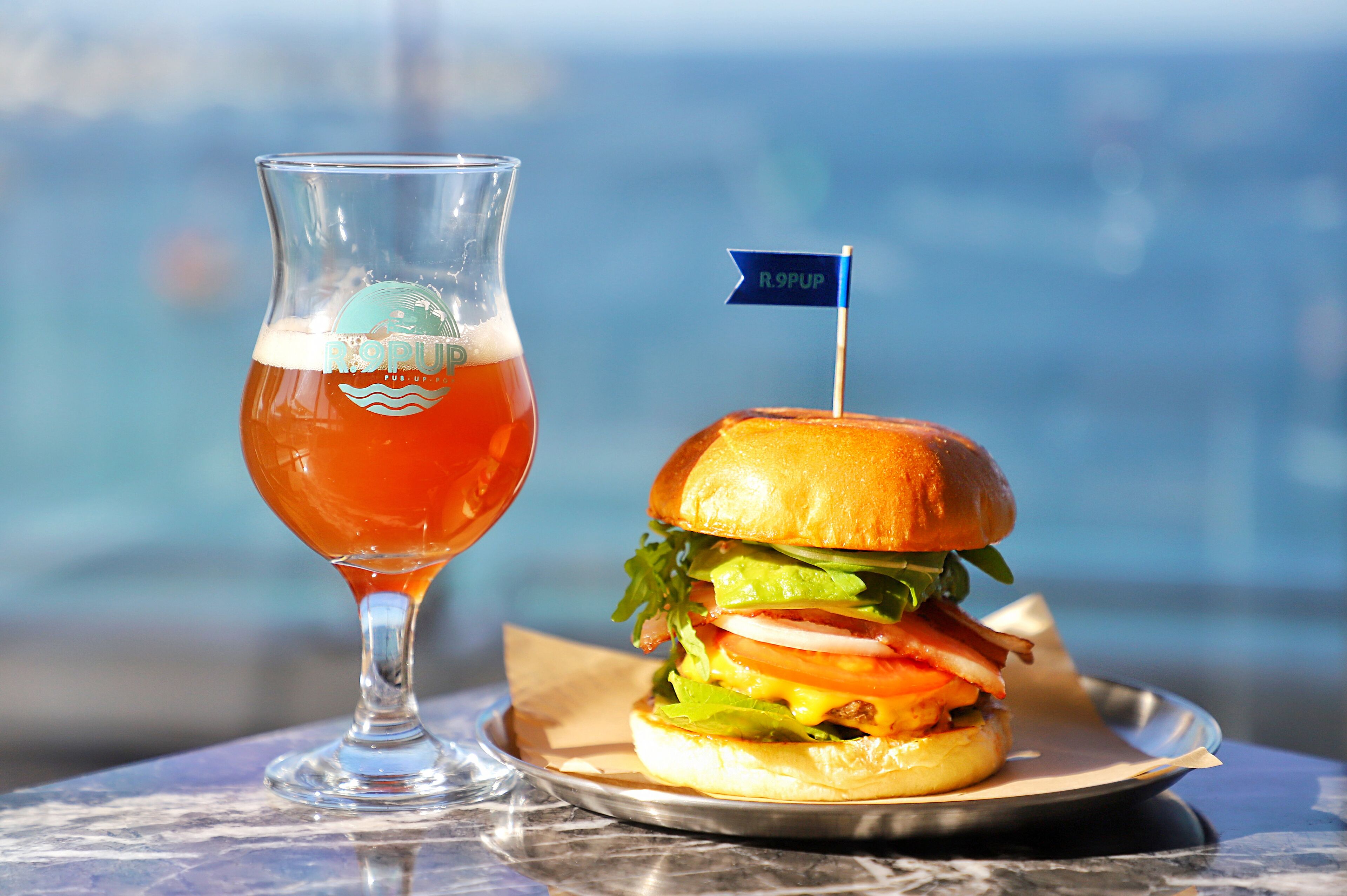 Una cerveza con una bandera sobre una mesa, junto a una hamburguesa.