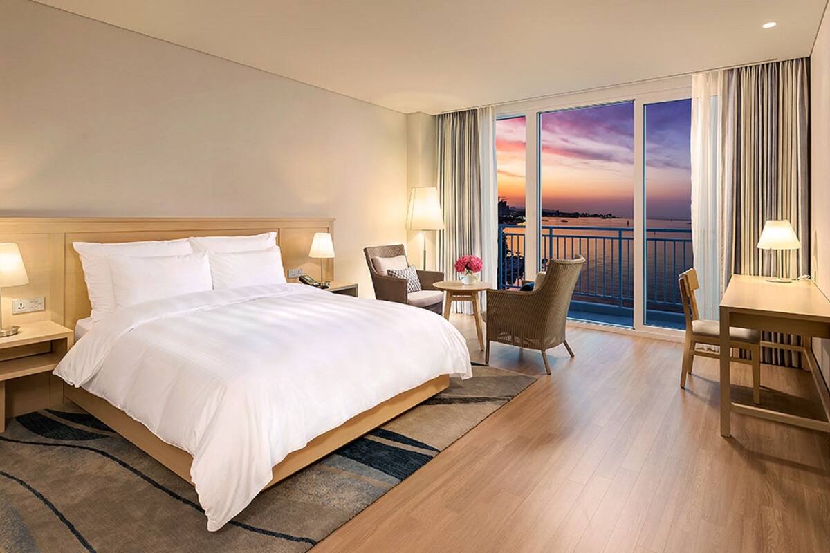 Une chambre d’hôtel avec un grand lit, deux tables de chevet avec des lampes, une chaise, un bureau et une vue sur le coucher de soleil à travers de grandes fenêtres.