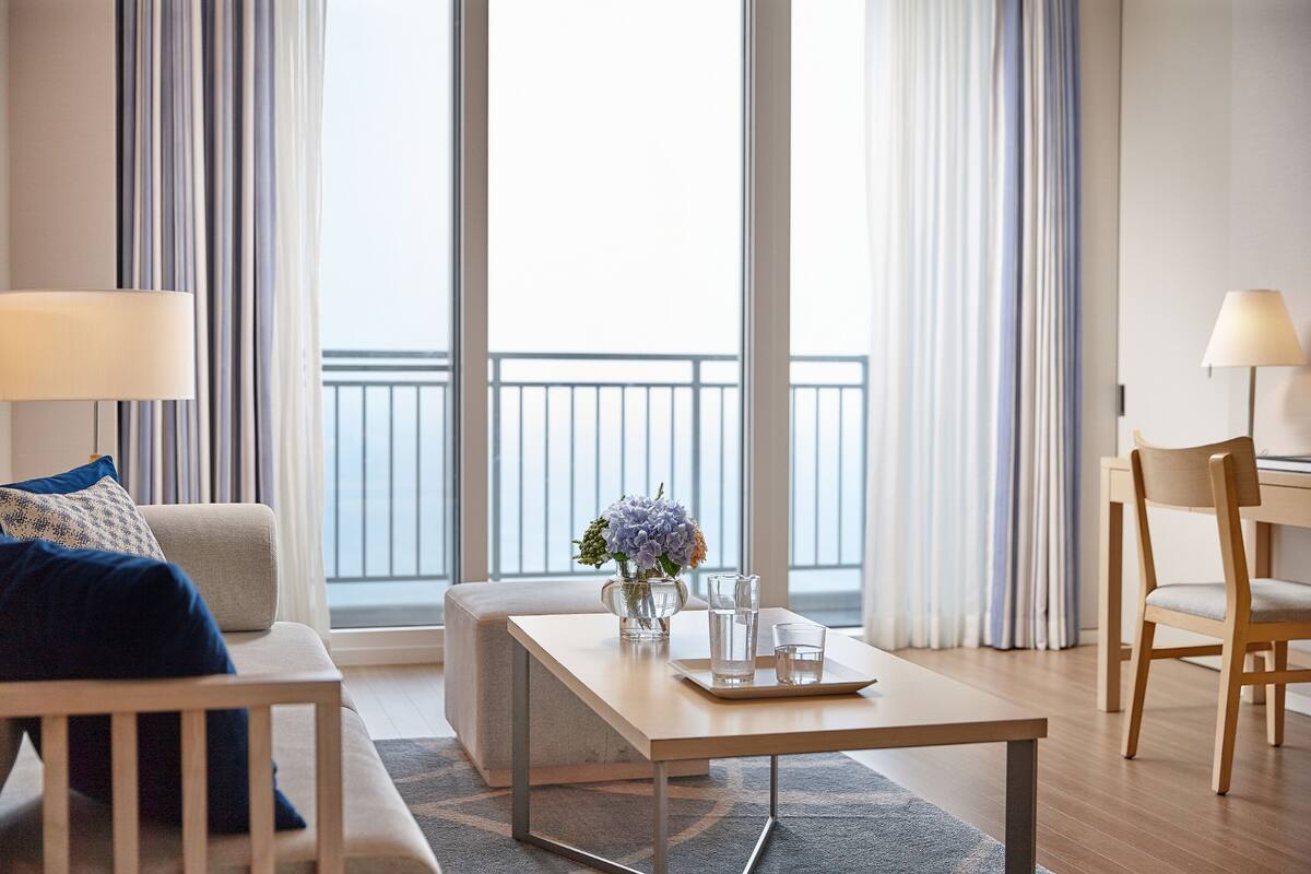 Une chambre d’hôtel moderne avec un canapé, une table basse agrémentée d’un vase et de verres, un fauteuil et une vue sur le balcon.