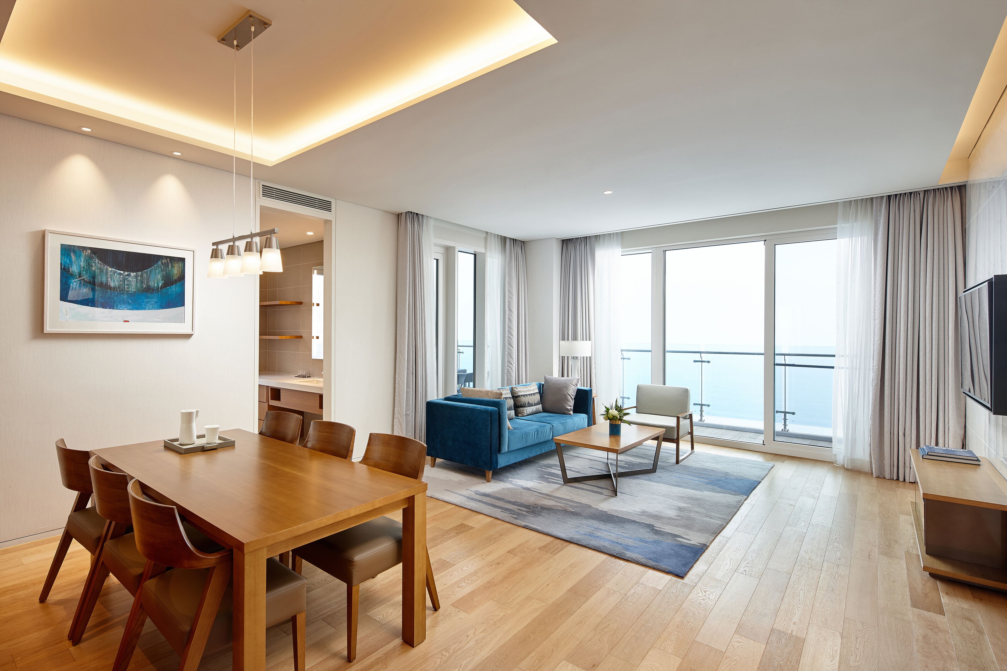A modern living room with a wooden dining table, blue sofa, and a large window with curtains.