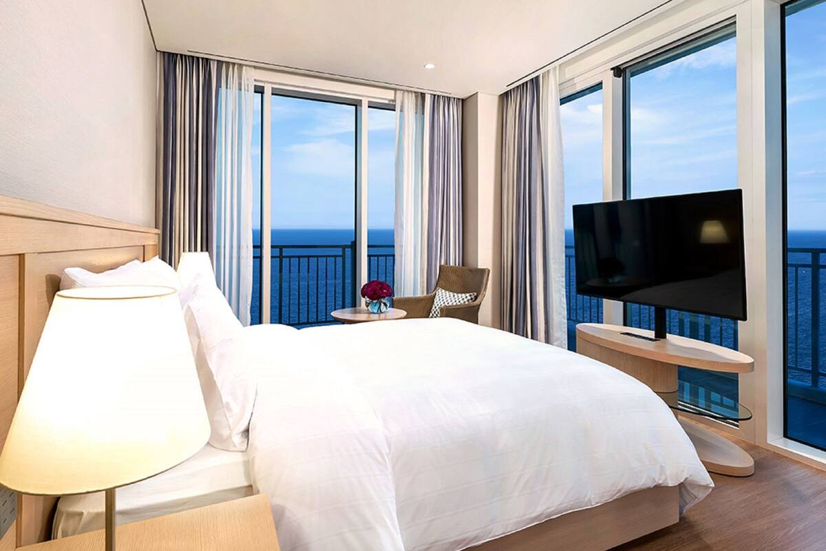 Une chambre d’hôtel avec un grand lit, une télévision, une petite table avec un vase et un balcon donnant sur la mer.