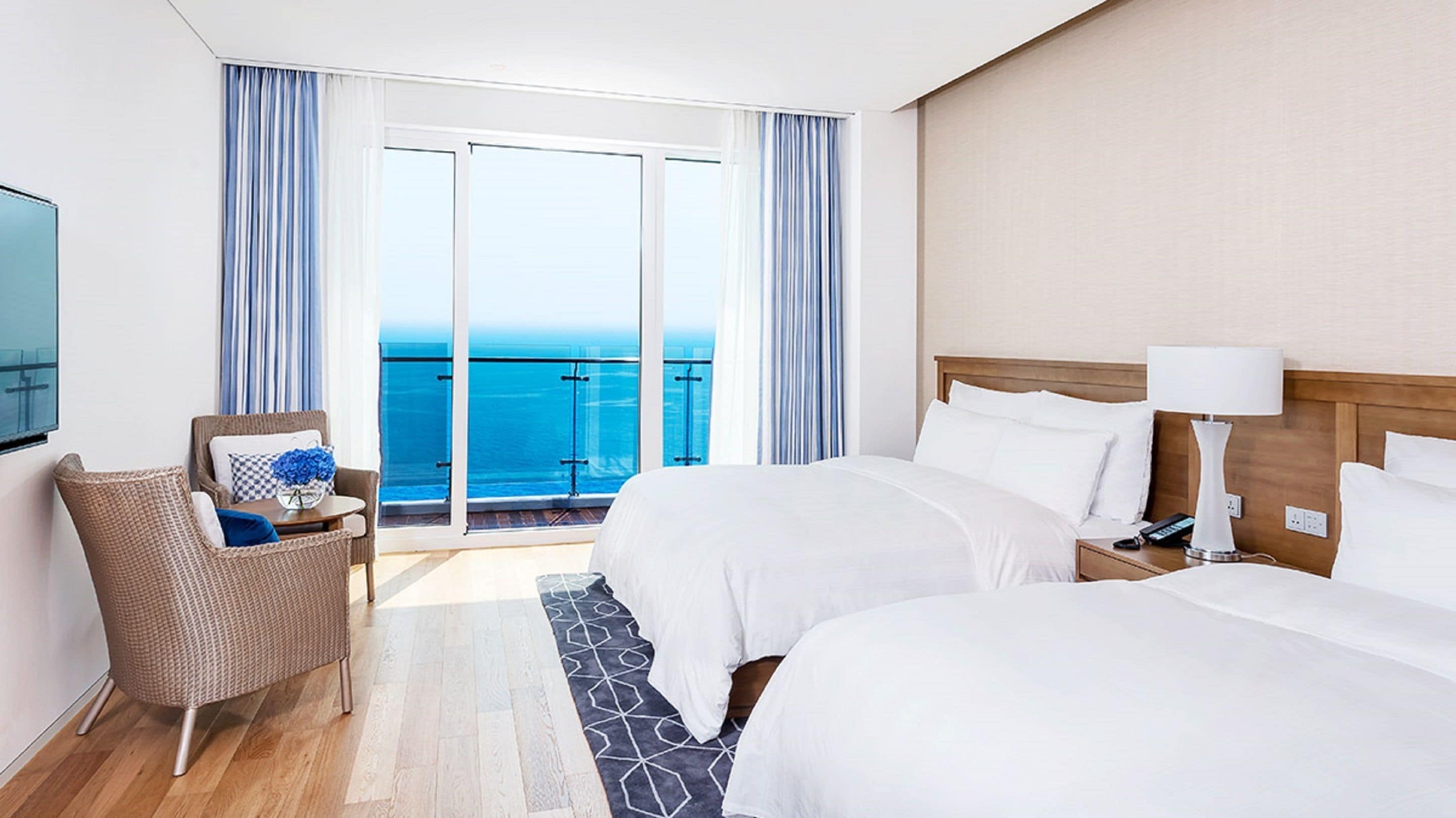 Une chambre d’hôtel avec deux lits, une chaise, une table ornée d’un vase de fleurs, une télévision et un balcon donnant sur la mer.