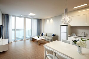 A modern living room with a sofa, coffee table, and dining table. There is a kitchen area with a sink and cabinets. A balcony with curtains is visible through the large windows.