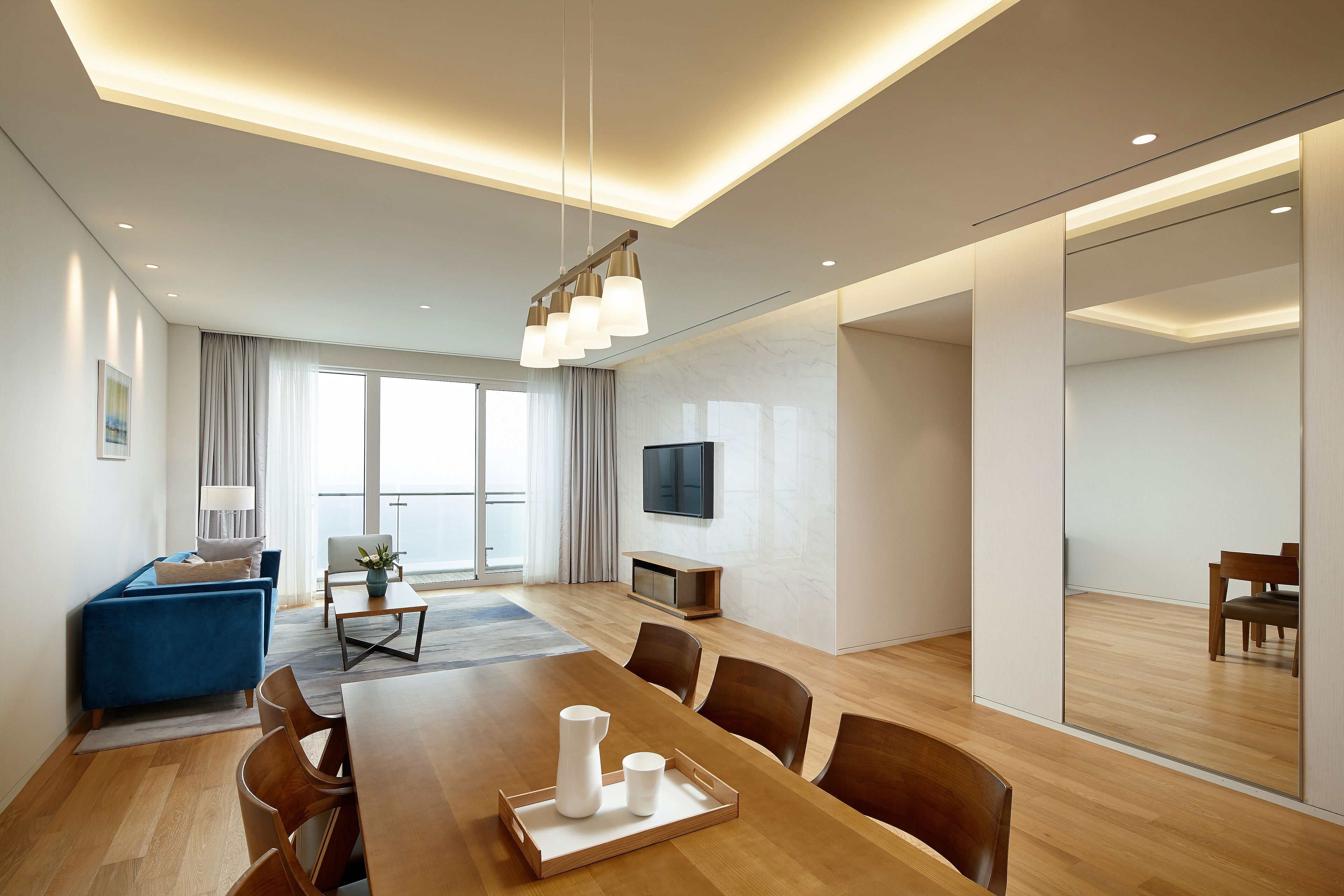 A modern hotel room with a dining area, a sofa, a TV, and a large window.