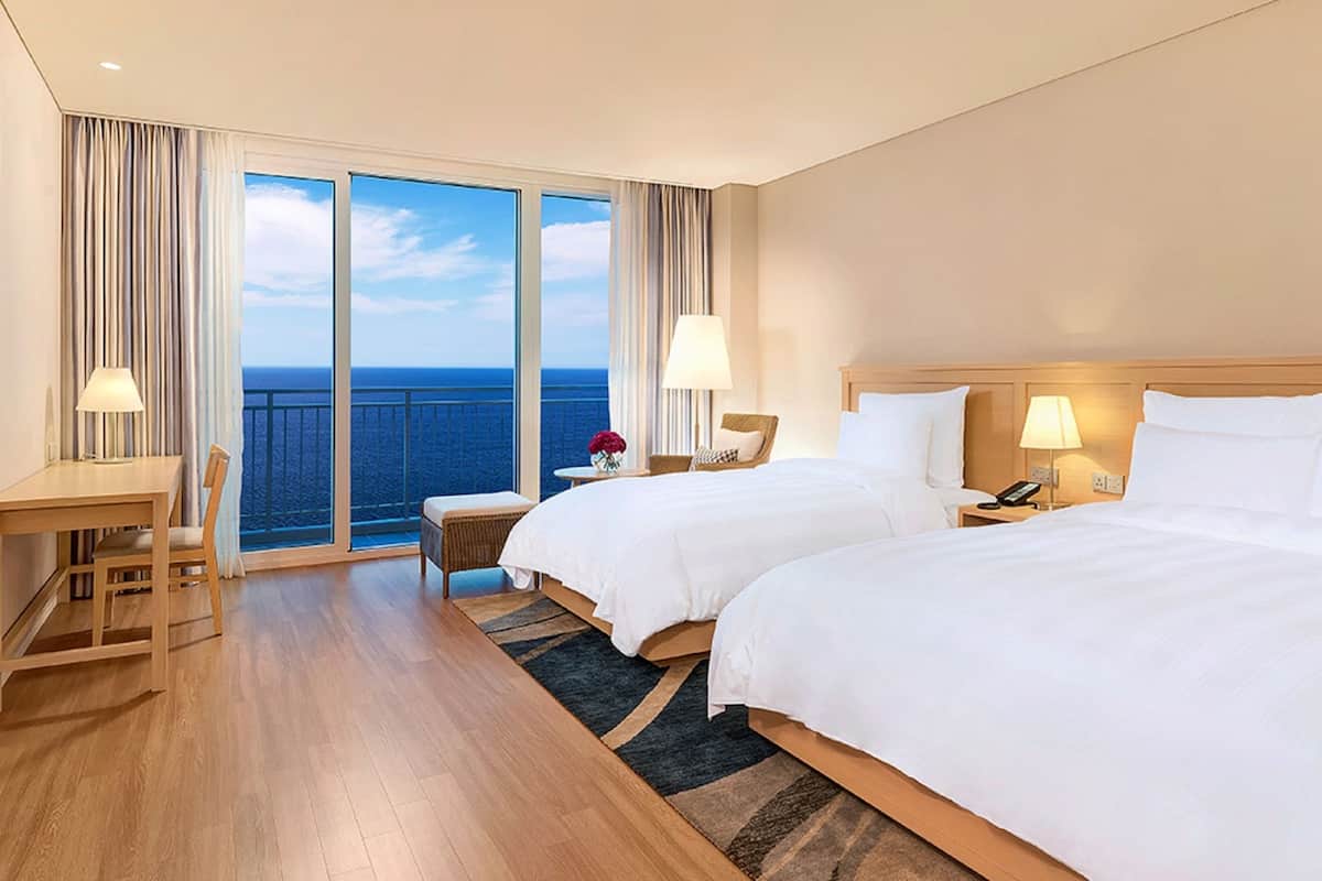 Une chambre d’hôtel avec deux lits, un bureau, une chaise, une petite table avec une lampe et une vue sur l’océan.