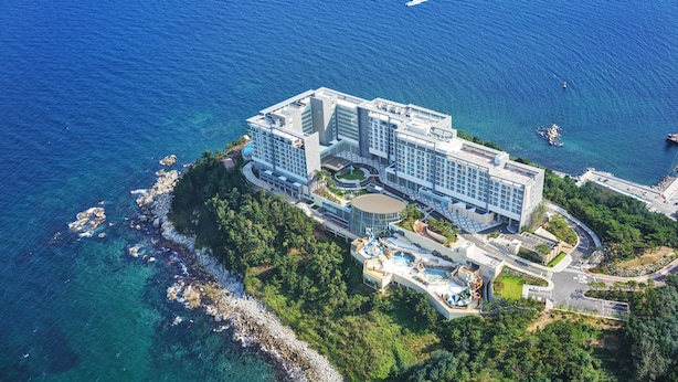 An aerial view of a coastal hotel with multiple buildings, a marina, and lush greenery. - Lotte Resort Sokcho (Sokcho)