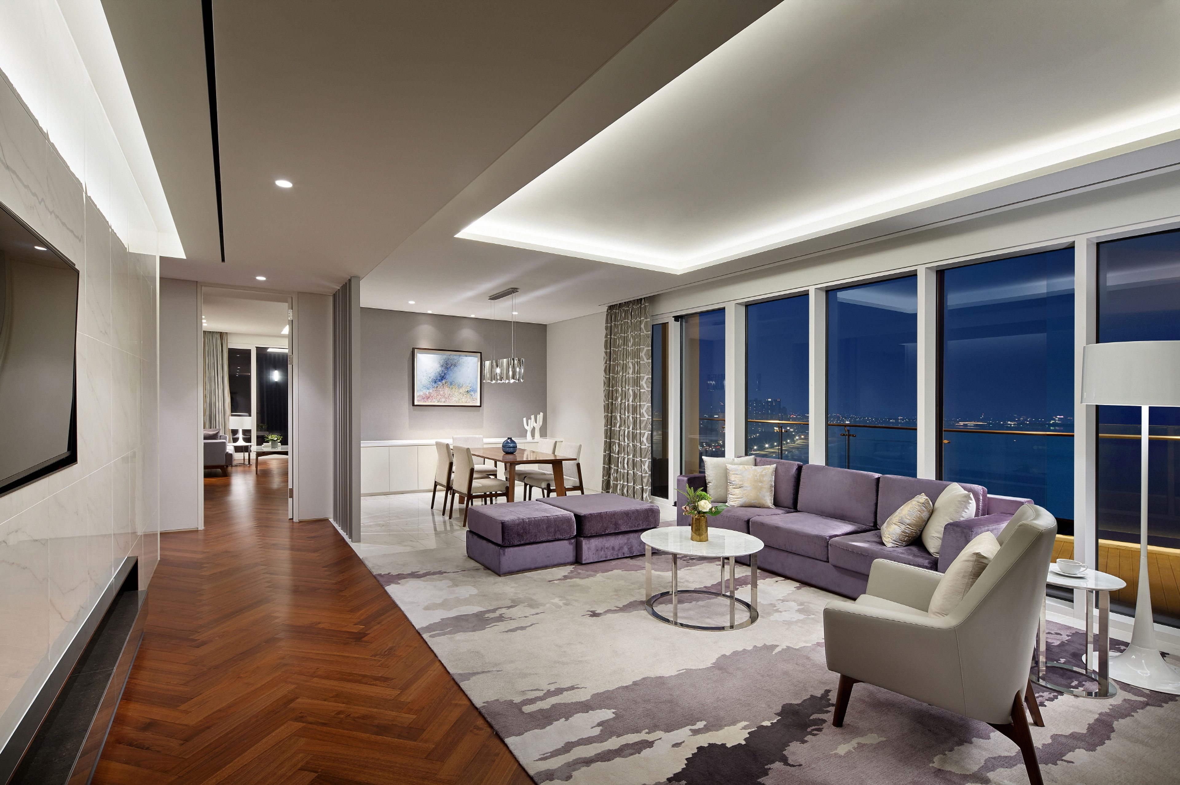 A modern living room with a sofa, armchair, and coffee table, a dining area with chairs and a table, and large windows with a city view.
