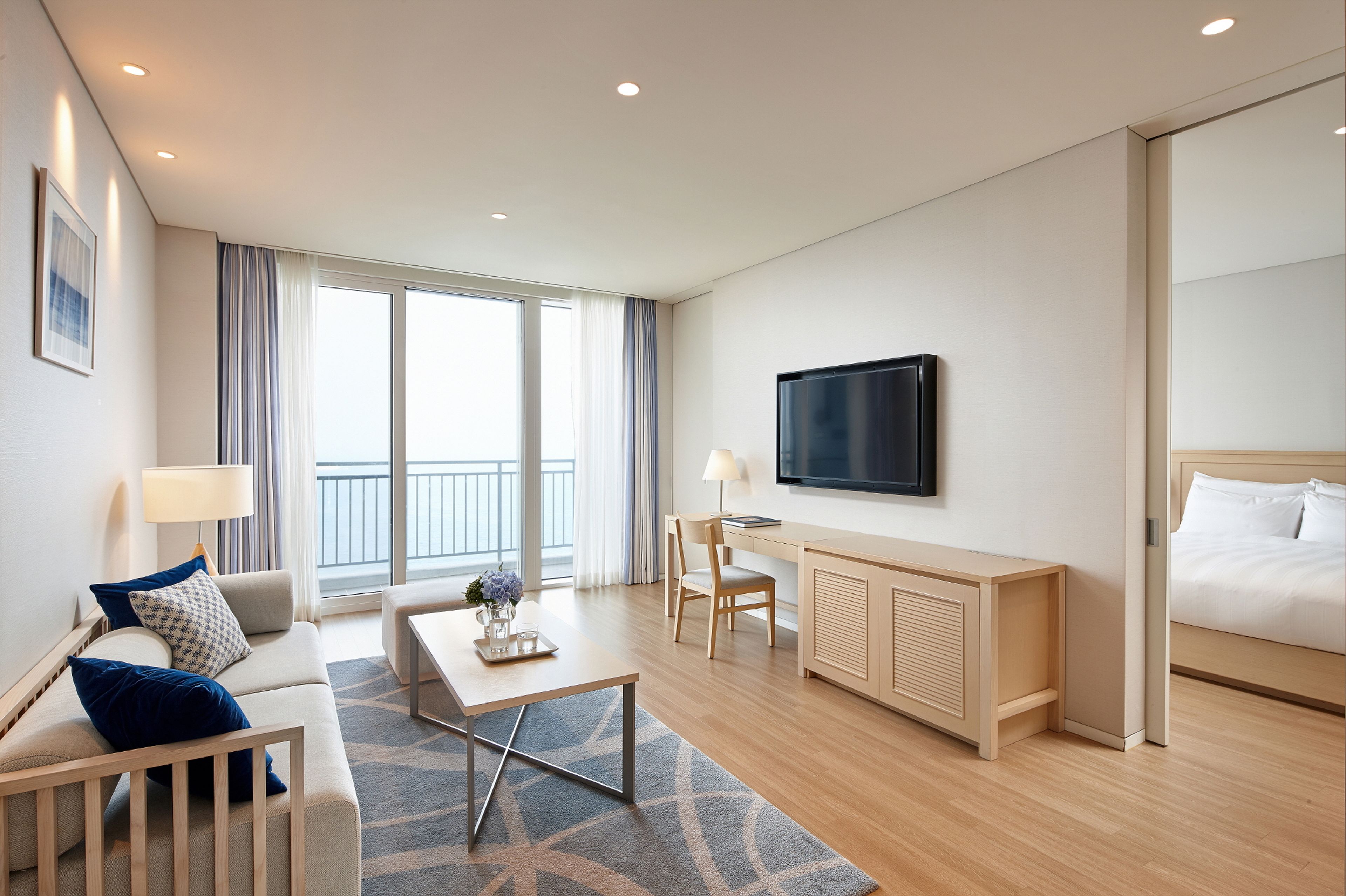 A modern hotel room with a sofa, a desk, a bed, a TV, and a balcony.