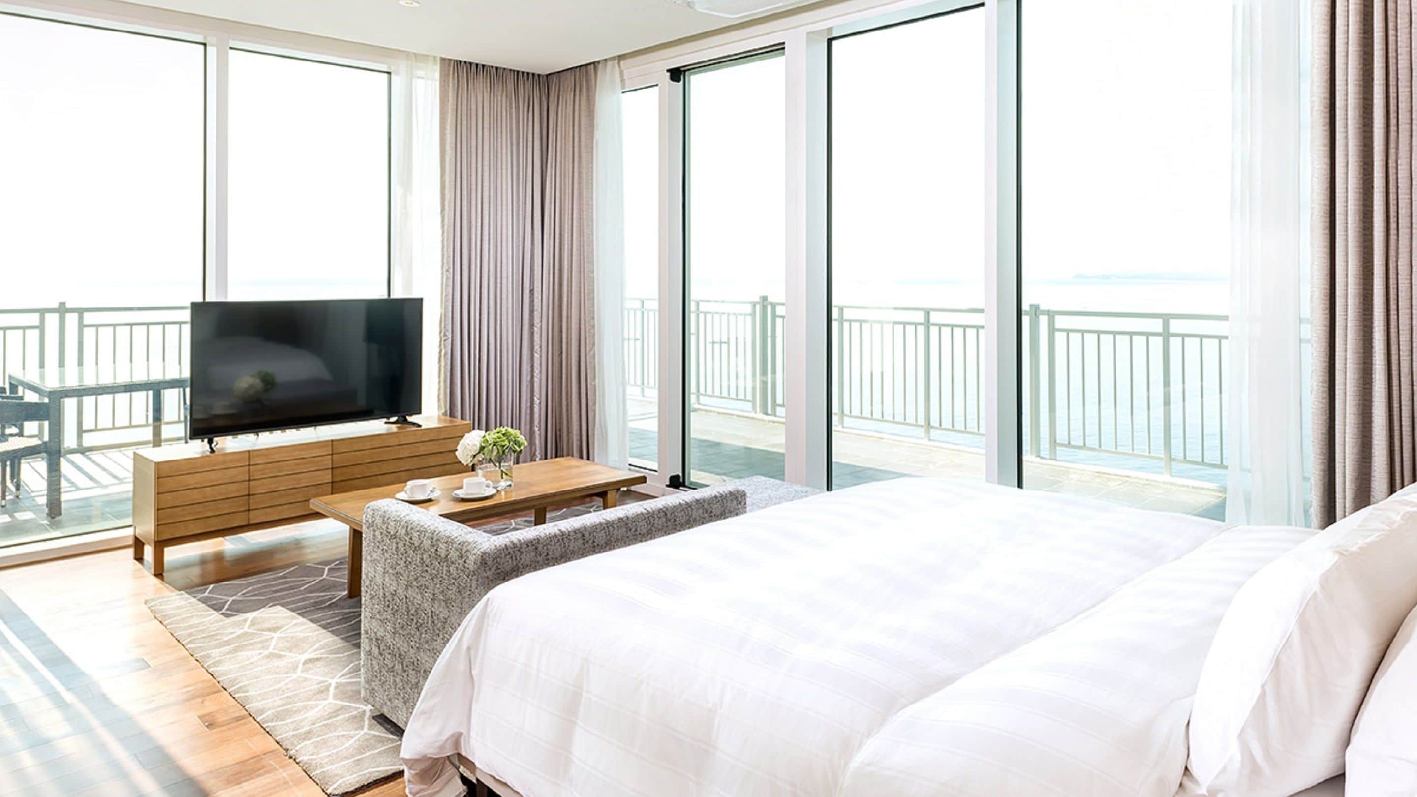 Et hotelværelse med en stor seng, et fjernsyn på et træpanel, en balkon med havudsigt og et siddeområde med et bord og stole.