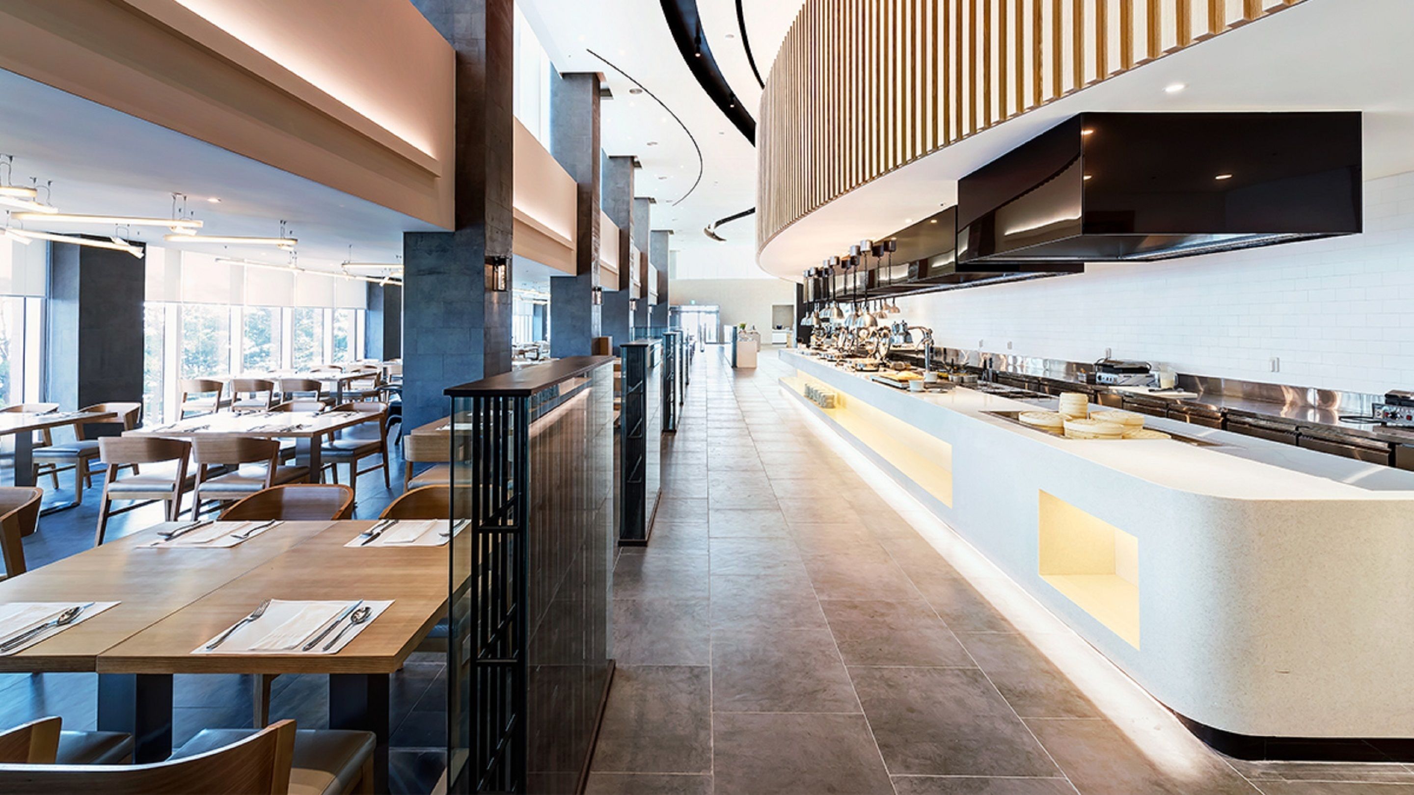 A modern restaurant with long communal tables, a central counter with a kitchen, and a high ceiling with exposed beams.