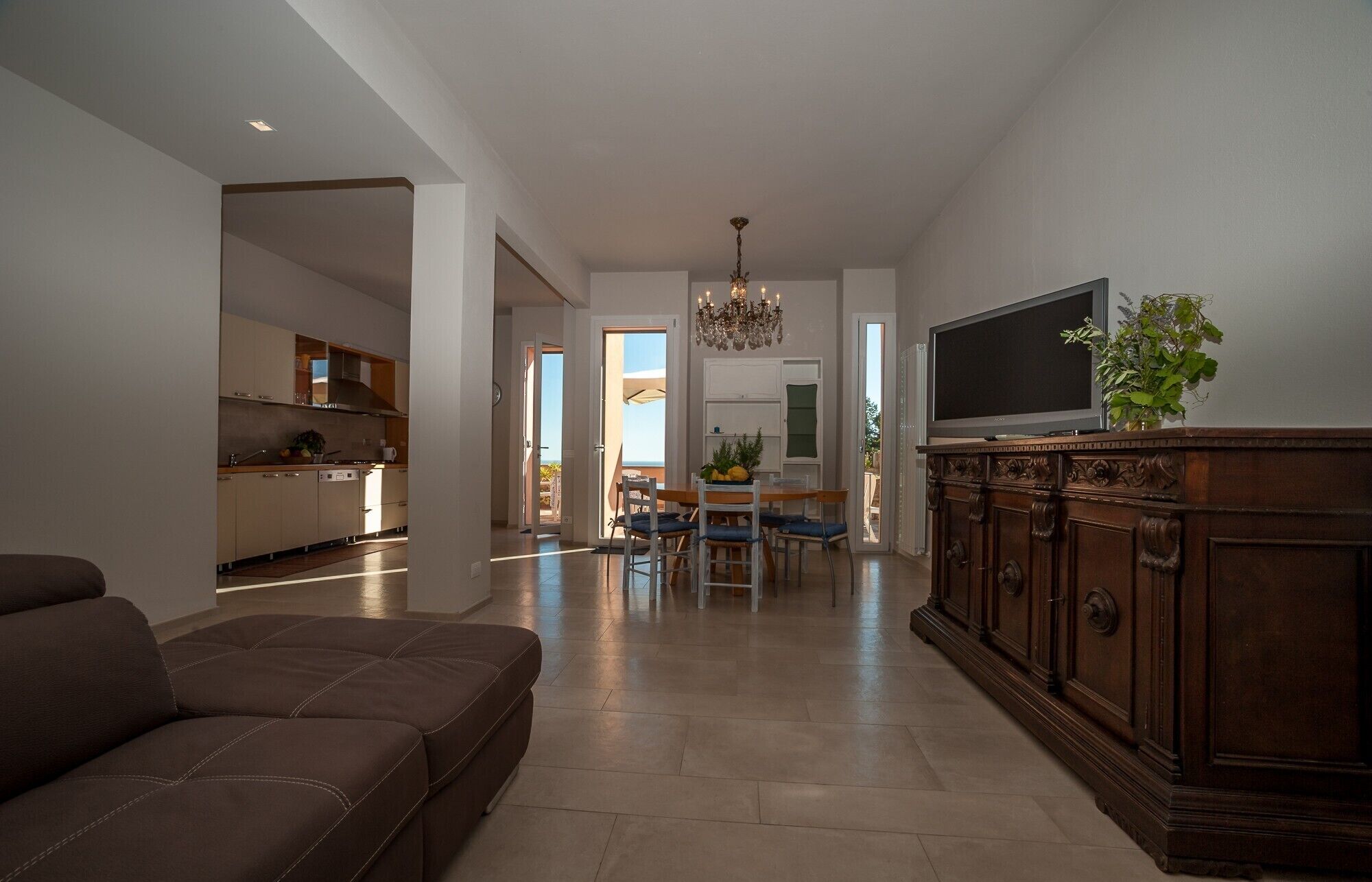 Un soggiorno moderno con un divano marrone, un'area pranzo con tavolo e sedie, una cucina con pensili e un piano cottura, e un televisore su un mobile in legno.