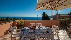 Una mesa redonda con mantel floral, rodeada de sillas blancas, ubicada en una terraza con vistas al mar y a las montañas.