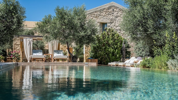 A clear swimming pool with lounge chairs and a pergola, surrounded by trees and a stone building.