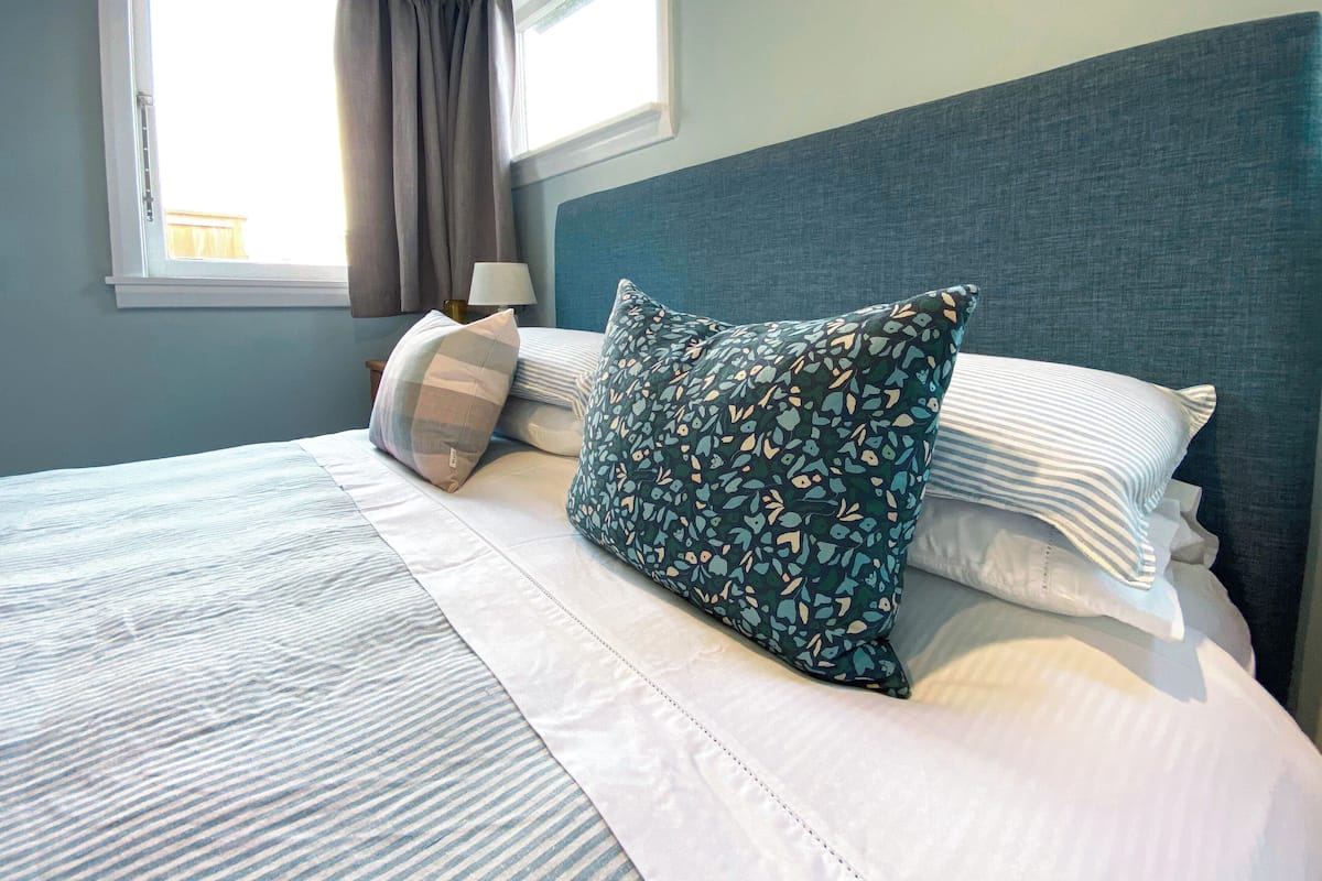 A neatly made bed with striped bedding and multiple pillows, including a floral-patterned one, in a room with a window and a lamp on a nightstand.