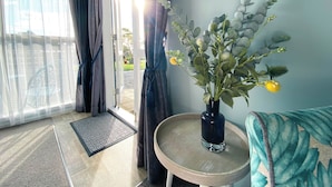 A room with a sliding glass door, a small round table with a vase of flowers, and a patterned sofa. - Kauri Villas (Parua Bay)