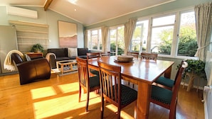 A spacious living room with a wooden dining table, comfortable seating, and large windows offering a view of greenery. - Kauri Villas (Parua Bay)