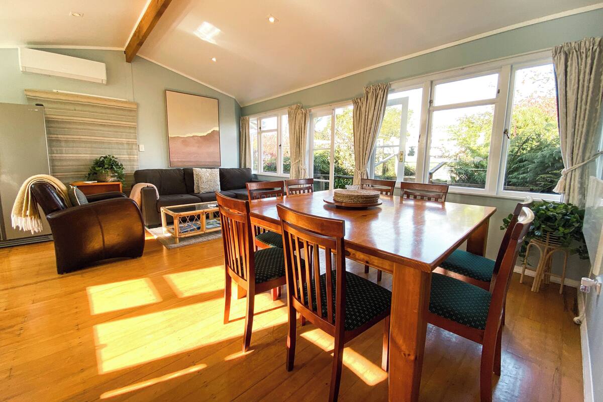 A spacious living room with a wooden dining table, comfortable seating, and large windows offering a view of greenery.