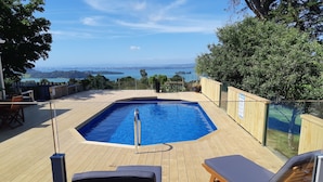A wooden deck with a swimming pool, lounge chairs, and a view of the sea. - Kauri Villas (Parua Bay)
