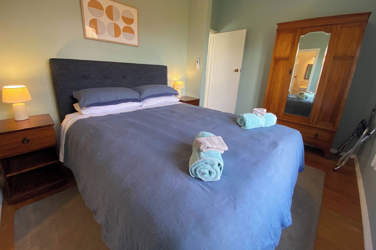 A neatly made bed with a blue bedspread and pillows, a wooden nightstand with a lamp, a wooden wardrobe, and a mirror reflecting part of the room.