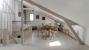 Ruime zolderkamer met hoge plafonds, zichtbare houten balken, een eethoek met rieten stoelen en een trap naar een ander niveau.