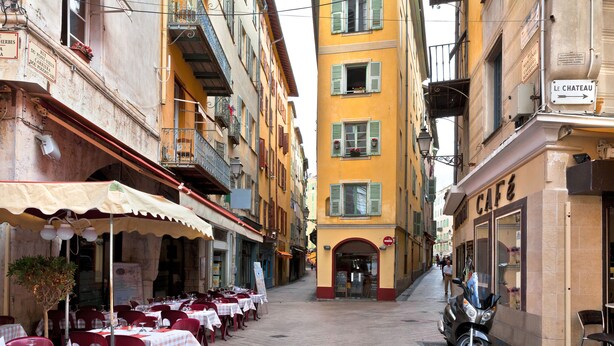 A narrow street lined with outdoor dining tables and chairs, a parked motorcycle, and a building with a sign reading "LE CHATEAU". - Air-conditioned studio with a garage near the Port of Nice (Nice)