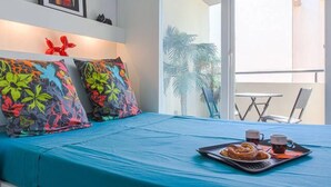 A hotel room with a bed, colorful tropical-themed pillows, a tray with pastries and drinks, and a view of the balcony with chairs and plants. - Air-conditioned studio with a garage near the Port of Nice (Nice)