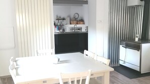 A dining area with a white table and chairs, a kitchen in the background, and a modern light fixture above the table. - Renovated stone house private next to river, Welcoming and very well equipped (Juvinas)