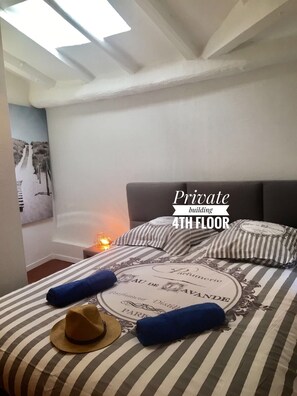 A bedroom with a bed featuring striped bedding, a hat, and a pillow. The room has a skylight and a wall with a painting. Text on the wall reads "Pratique building 4TH FLOOR." - PRIVATE BUILDING, 4 APTS, AIR CONDITIONING, WIFI - UNIQUE IN NICE - OLD NICE (Nice)