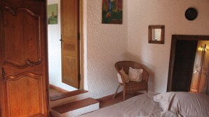 A bedroom with a wooden wardrobe, a wicker chair, a bed, and a door leading to another room.