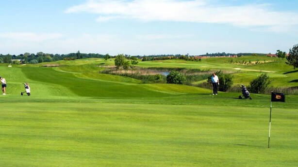 En golfbana med böljande, grönskande kullar, en damm och en flaggstång.