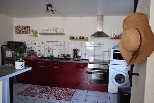 A compact kitchen with a microwave, stove, and washing machine. There is a potted plant and a decorative hat on the counter.