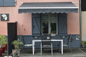 Une terrasse avec une table et des chaises, une fenêtre avec des volets et des plantes en pot.