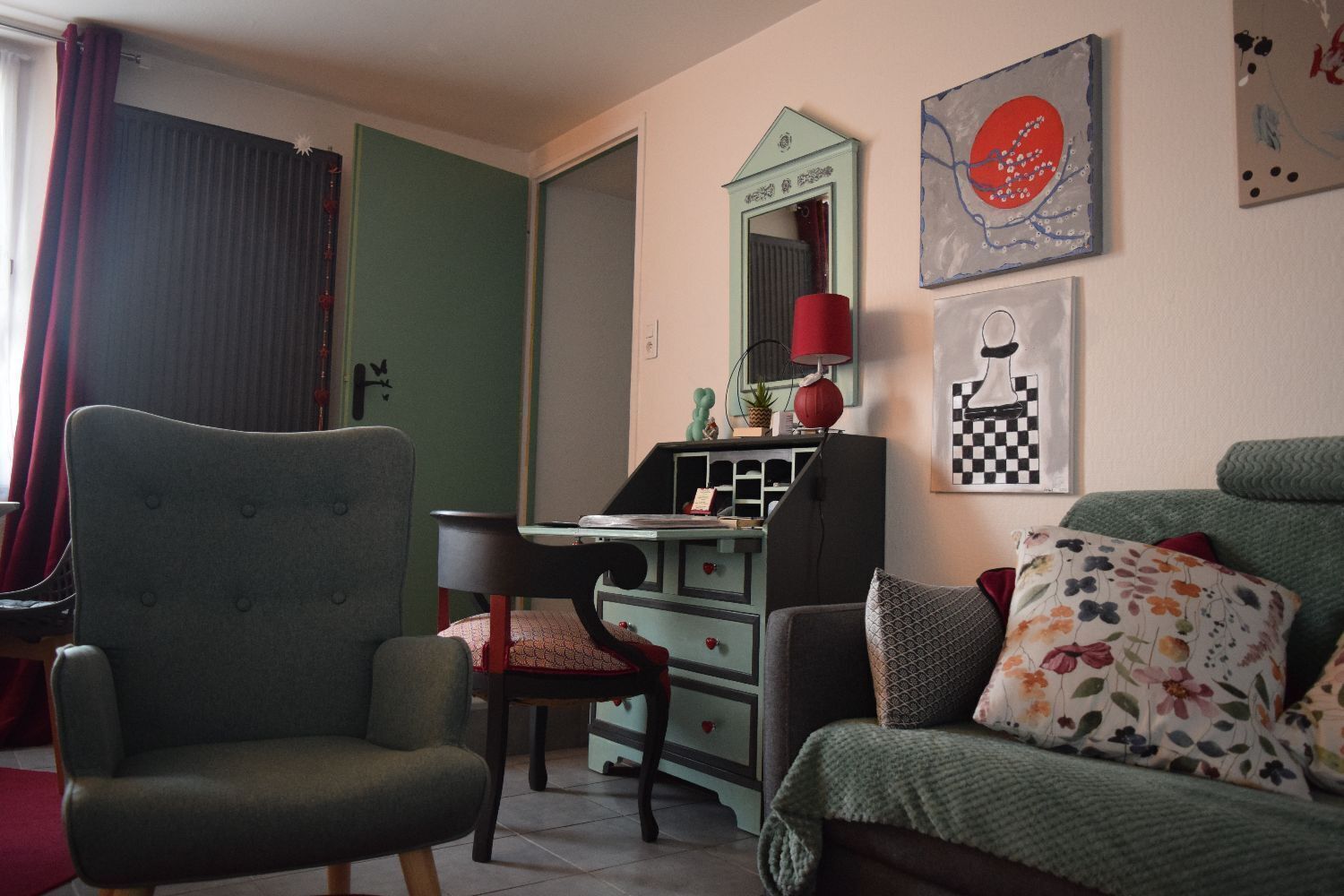 A cozy room with a green armchair, a wooden chair, a desk with a lamp, and a bed with floral pillows.
