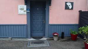 L’entrée d’un immeuble, avec une porte bleue, une enseigne indiquant « GITE LE GINKO » et deux plantes en pot posées sur le sol.