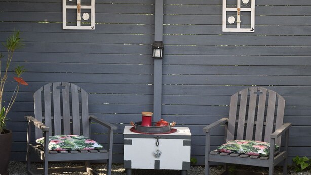 Deux fauteuils Adirondack avec des coussins à motifs floraux, une petite table avec une tasse rouge et un luminaire fixé au mur.