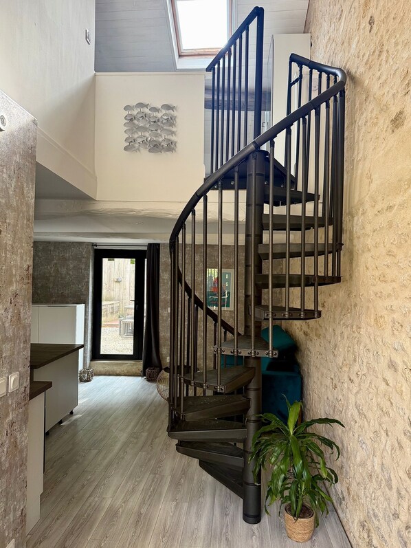 Un escalier en colimaçon avec une rampe en métal, un pot de fleurs et une vue sur l’espace extérieur à travers une porte vitrée.