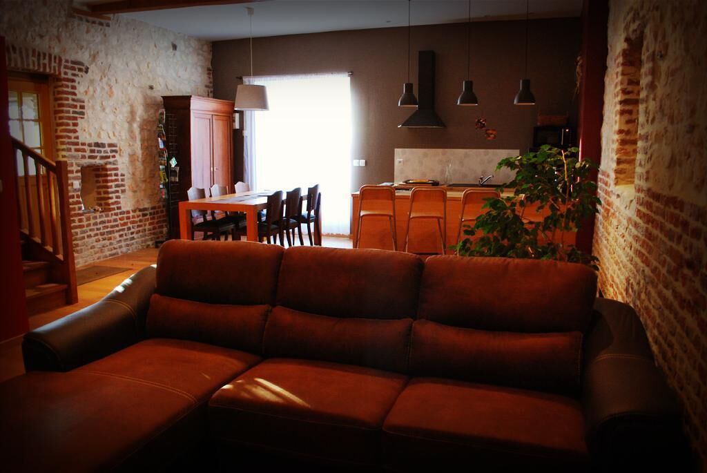 A cozy living room with a leather sofa, a dining area with a wooden table and chairs, and a kitchen area with a countertop and hanging lights.