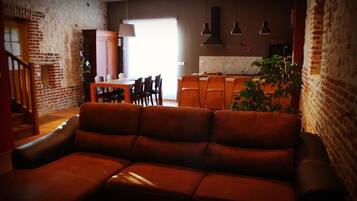 A cozy living room with a leather sofa, a dining area with a wooden table and chairs, and a kitchen area with a countertop and hanging lights.