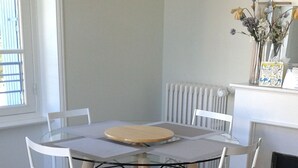 Zona pranzo con tavolo rotondo, sedie bianche, tappeto nero e giallo e una finestra con persiane.